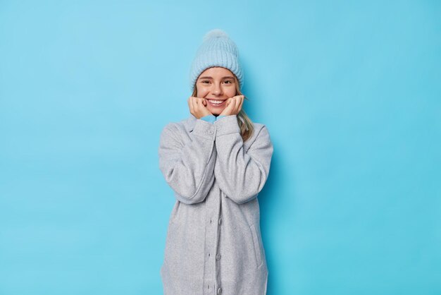 Binnenopname van vrij millennial Europees meisje glimlacht zachtjes houdt handen onder kin draagt grijze jas en hoed hoort aangenaam nieuws geïsoleerd over blauwe studio achtergrond. Gelukkig emoties concept