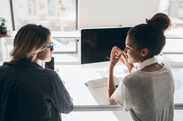 Binnenportret van achterkant van twee vrouwen die achter computer met zwart scherm zitten. Afrikaanse vrouwelijke beambte in koptelefoon praten met collega naast venster.