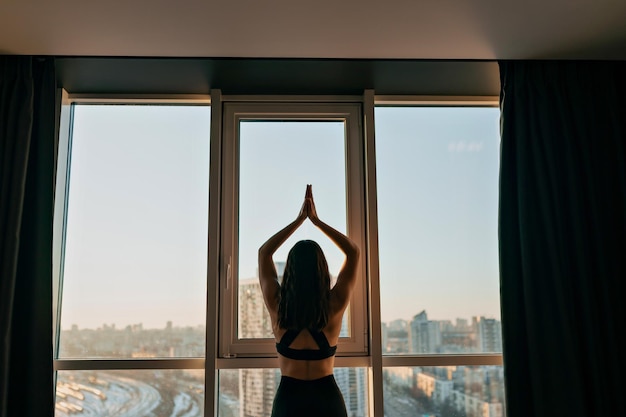 Binnenportret van de achterkant van een jonge, fitte vrouw in uniform met vlekken doet yoga met uitzicht op de stad in de ochtendzon