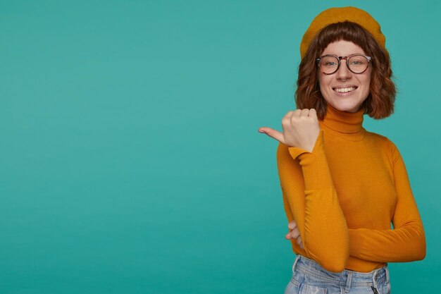 Binnenportret van een jonge gembervrouw draagt een oranje sweatshirt en een bril die over een blauwe achtergrondpunt poseert met een vinger opzij en glimlacht Gratis Foto