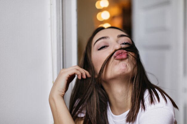 Binnenportret van gelukkig vrouwelijk model dat gezichten maakt. Foto van speelse bruinharige dame poseren met kussende gezichtsuitdrukking.