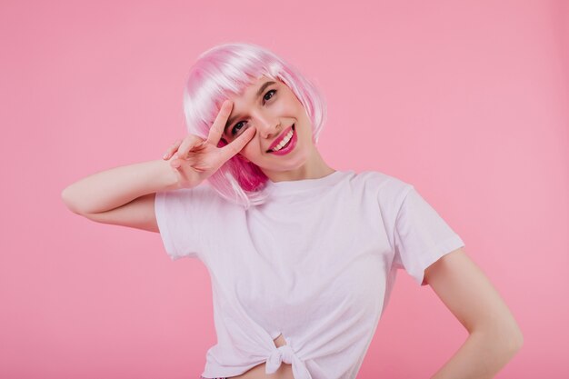 Binnenportret van glimlachend mooi meisje met roze haar dat op pastelkleurmuur wordt geïsoleerd. bevallige blanke dame in wit t-shirt poseren met vredesteken en lachen
