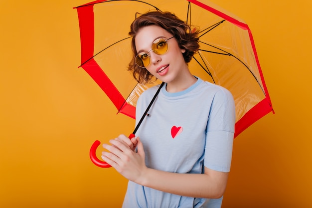 Binnenportret van mooi meisje in de blauwe parasol van de overhemdsholding. Foto van prachtige blanke dame met krullend kapsel geïsoleerd op gele muur met paraplu.