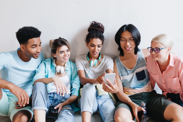 Binnenportret van vrolijke studenten die hun telefoons houden en glimlachen. Sierlijk Afrikaans meisje in oortelefoons en spijkerbroek maken selfie met vrienden op de universiteit.