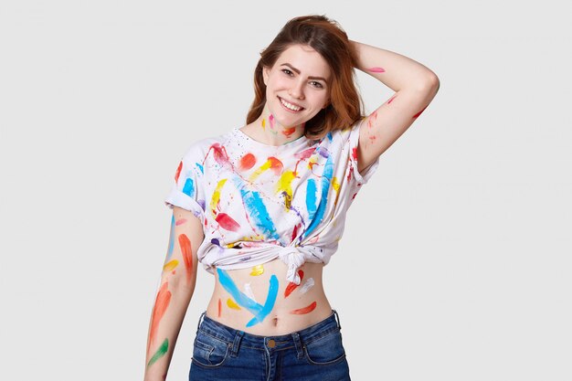 Binnenschot van gelukkige Europese vrouwelijke schilder heeft een vies lichaam en een wit t-shirt met kleurrijke verf, toont buik, houdt hand achter hoofd, geïsoleerd over witte muurcreaties, kunstwerk of meesterwerk