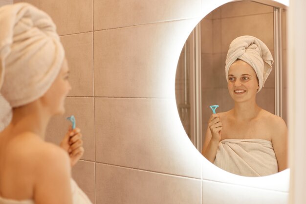 Binnenschot van gelukkige jonge volwassen vrouw met scheermes voor het scheren van de oksel in de hand, poseren in de badkamer voor ontharing, kijken naar spiegelreflectie, hygiëneprocedures.