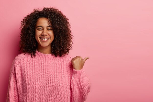 Binnenshuis shot van tevreden Afro-vrouw heeft donker krullend haar, wijst met duim opzij, toont mooie kopie ruimte voor uw reclame, heeft vriendelijke uitdrukking, draagt een oversized roze trui.