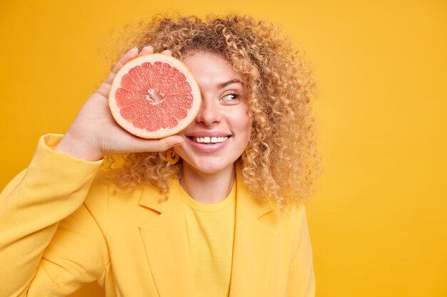 Biologisch dieet concept. Blije vrouw met krullend haar bedekt oog met grapefruit halve glimlacht tandjes die vers sap of smoothie gaan maken kijkt opzij poses tegen gele muur kopieerruimte voor tekst