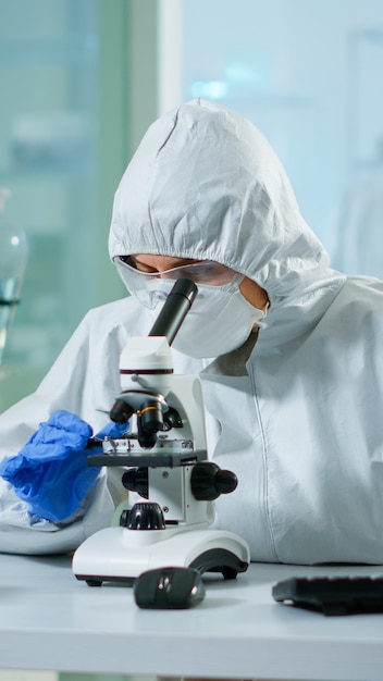 Bioloog arts in overall die DNA-monster controleert op microscoop typen op pc in uitgerust lab. Onderzoek naar de evolutie van vaccins in een medisch laboratorium met behulp van hightech- en scheikundige hulpmiddelen voor wetenschappelijk onderzoek