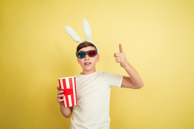 Bioscoop kijken in een bril met popcorn. Blanke jongen als paashaas op gele achtergrond. Gelukkig Pasen. Prachtig mannelijk model. Concept van menselijke emoties, gezichtsuitdrukking, vakantie. Copyspace.