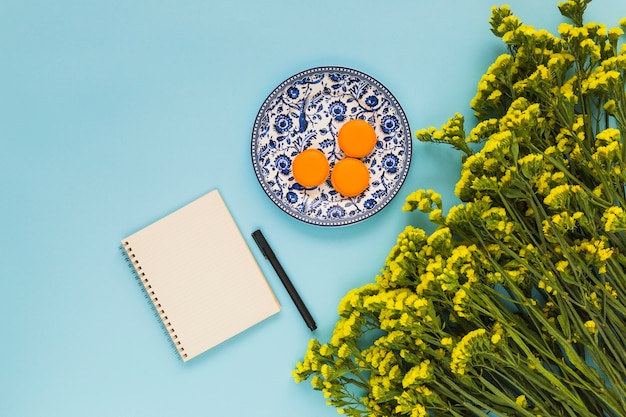 Bitterkoekjes op keramische plaat; spiraal notitieblok; pen en stelletje gele bloemen op blauwe achtergrond