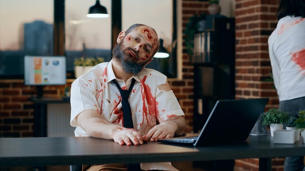 Bizar uitziende zombie met behulp van moderne laptop in kantoorwerkruimte. kwaad apocalyptisch hersenetend monster met diepe en bloedige wonden en littekens die een draagbare computer op het werk proberen te gebruiken.