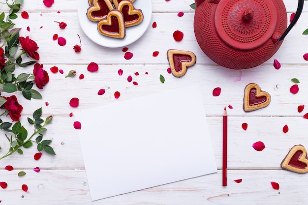 Blanco papier met rode rozenblaadjes op een wit oppervlak in de buurt van een rode theepot en hartvormige koekjes
