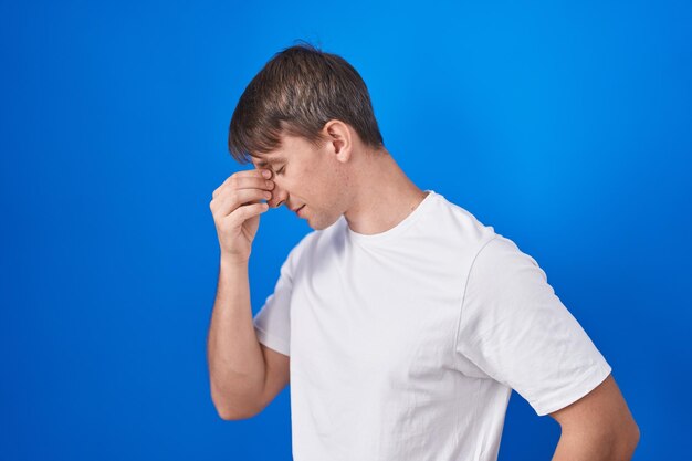 Blanke blonde man staande over blauwe achtergrond moe wrijven neus en ogen vermoeidheid en hoofdpijn voelen. stress en frustratie concept.