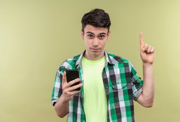 blanke jonge man met een groen shirt met telefoon wijst omhoog op geïsoleerde groene muur