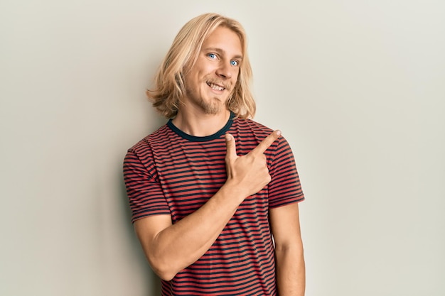 Gratis foto blanke jonge man met lang haar, gekleed in casual gestreept t-shirt glimlachend vrolijk wijzend met hand en vinger omhoog naar de zijkant