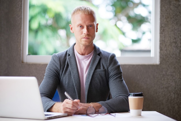 Blanke man in blazer zit aan bureau in kantoor met laptop en camera kijken