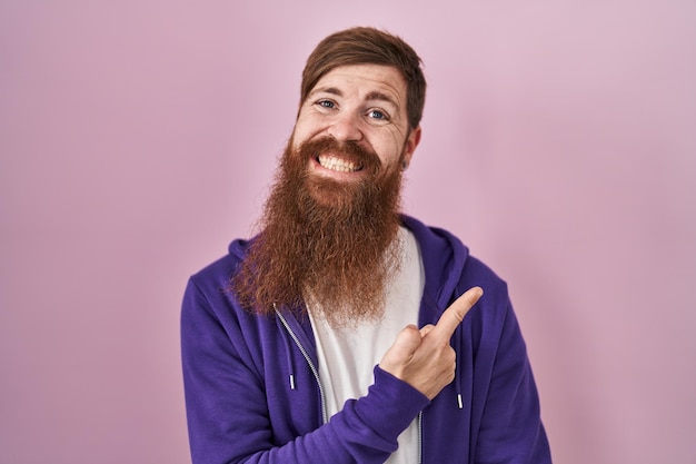 Gratis foto blanke man met een lange baard die over een roze achtergrond staat, vrolijk met een glimlach van het gezicht wijzend met hand en vinger naar de zijkant met een gelukkige en natuurlijke uitdrukking op het gezicht