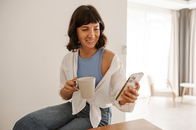 Blanke vrouw die met koffie naar de telefoon kijkt