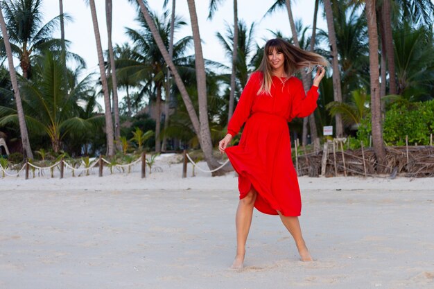 Blanke vrouw in rode zomerjurk in romantische gelukkige stemming op tropisch wit zandstrand bij zonsondergang