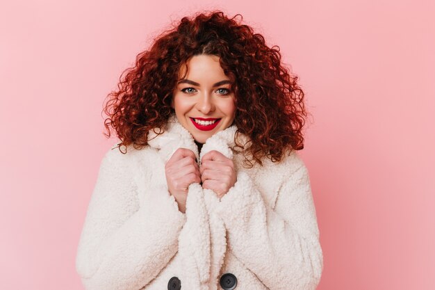 Blauwogige krullende meisje met rode lippenstift lacht en poseren in witte eco-jas op roze ruimte.