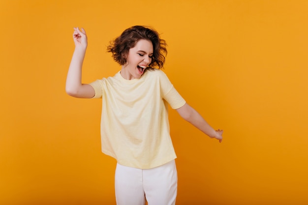 Bleek bruinharig meisje in geel t-shirt dat met geïnspireerde gezichtsuitdrukking danst. Actieve jonge vrouw in casual zomer outfit plezier indoor.