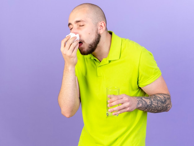 Gratis foto bleke jonge blanke zieke man veegt neus met weefsel af en houdt glas water op paars