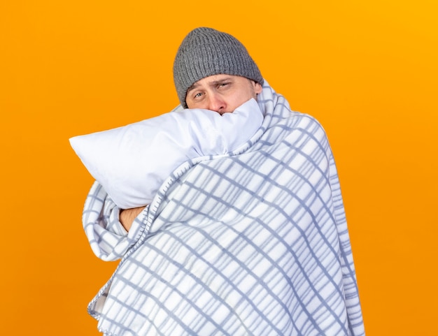 Bleke jonge blonde zieke man met winter hoed verpakt in geruite knuffels kussen geïsoleerd op een oranje muur