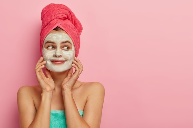 Blij, aangenaam uitziend vrouwelijk model heeft gezichtsmasker voor vochtige huid, toont schoon, fris gezicht, halfnaakt lichaam gewikkeld in handdoek, kijkt opzij op kopie ruimte