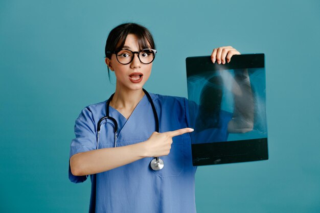 Blij bedrijf en wijst op xray jonge vrouwelijke arts dragen uniforme fith stethoscoop geïsoleerd op blauwe achtergrond