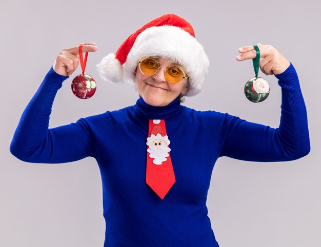 Blij bejaarde vrouw in zonnebril met kerstmuts en santa stropdas met glazen bol ornamenten geïsoleerd op een witte achtergrond met kopie ruimte