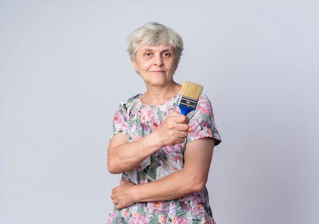 Blij bejaarde vrouw legt hand op buik houdt kwast geïsoleerd op een witte muur