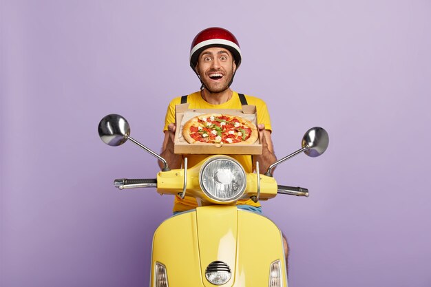 Blij bekwame bezorger die gele scooter bestuurt terwijl hij pizzadoos vasthoudt