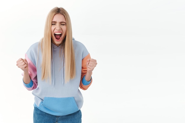 Blij blij overweldigd blond meisje winnen triomferend over succes balde vuisten en schreeuwen ja vieren overwinning bereiken doel winnen prijs en zingen staande witte achtergrond