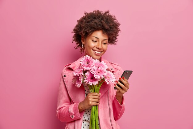 Blij dat Afro-Amerikaanse dame in stijlvolle kleding controleert berichten online krijgt gefeliciteerd met verjaardag houdt mooi boeket gerbera's heeft een gelukkige stemming geïsoleerd over roze muur