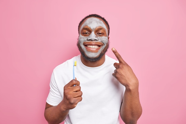 Blij dat Afro-Amerikaanse man glimlacht, geeft graag om tanden, houdt tandenborstel vast en brengt schoonheidskleimasker op gezicht aan