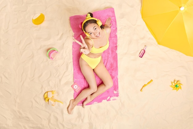 Blij dat Aziatische vrouw op het strand zit en vredesgebaar maakt rillingen tijdens de zomerdag zonnebaadt heeft plezier en luistert naar favoriete muziek via een koptelefoon gebruikt een parasol om zich te verbergen voor de zon brengt vakantie in het buitenland door