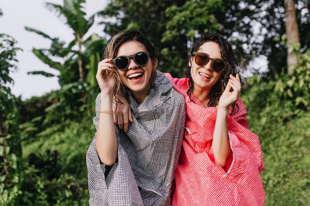 Blij dat donkerharige vrouwen in zonnebril lachen om de natuur. Goedgehumeurde vrouwelijke toeristen in regenjassen die plezier hebben in de jungle.