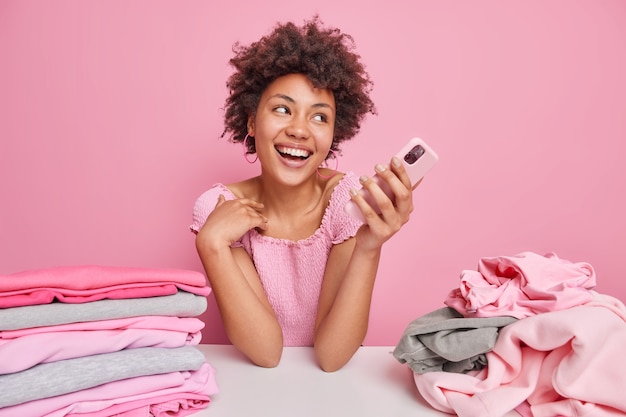 Blij dat een vrouw met een donkere huid met krullend haar een mobiele telefoon vasthoudt en wegkijkt heeft een vrolijke uitdrukking aan tafel met wasgoed in de buurt doet huishoudelijke klusjes geïsoleerd over roze