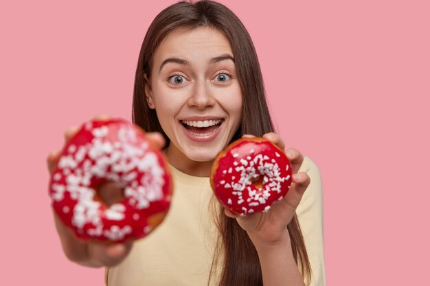 Blij dat gelukkige jonge vrouw een brede glimlach heeft, in een hoge geest is, een smakelijk dessert draagt, zich op douchnuts richt, vrijetijdskleding draagt, geïsoleerd op roze achtergrond. Mensen