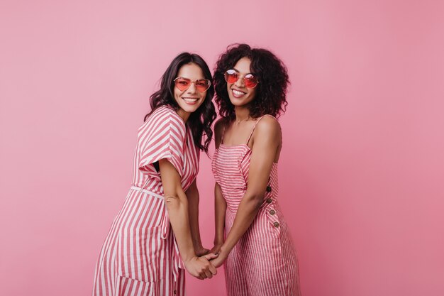 Blij dat jonge dames elkaars hand vasthouden tijdens het poseren. Binnen schot van lachende gelukkige vrouwelijke modellen draagt zomerkleding.