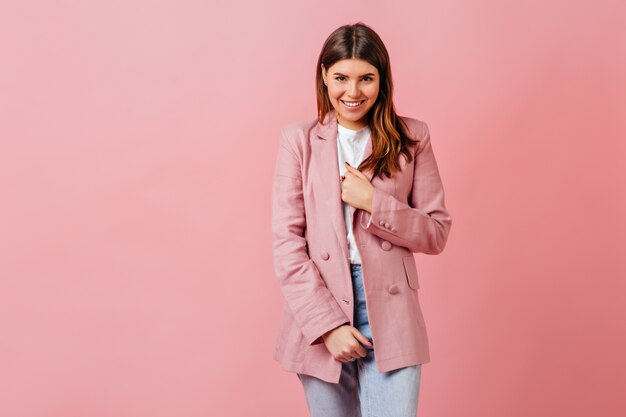 Blij dat jonge vrouw in jasje bij camera glimlacht. Studio shot van aantrekkelijke brunette dame geïsoleerd op roze achtergrond.