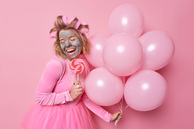 Blij dat jonge vrouw klei schoonheidsmasker aanbrengt voor huidbehandeling houdt karamel snoep op stok en bos van opgeblazen helium ballonnen draagt feestelijke jurk maakt kapsel geïsoleerd over roze achtergrond