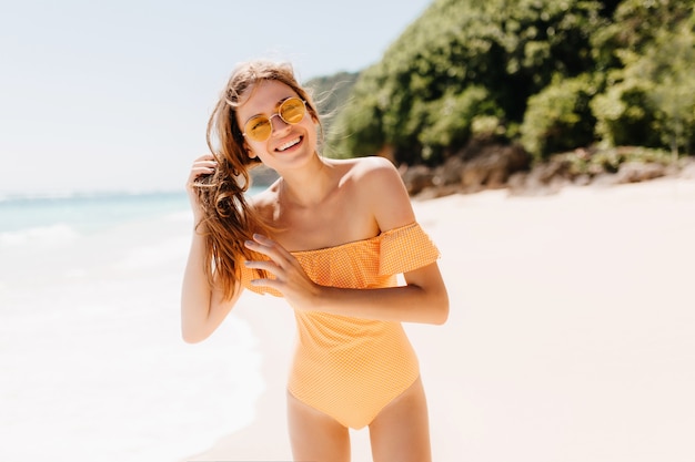 Blij dat sportieve vrouw haar bruine haar aanraakt tijdens het poseren op zandstrand. Betoverend Kaukasisch meisje in gele badmode gek rond op tropisch eiland.