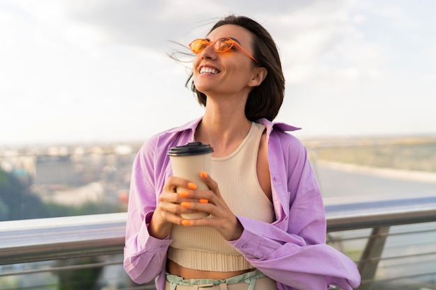 Blij dat vrouw met kort haar in stijlvolle zomeroutfit koffie drinkt op de moderne brug