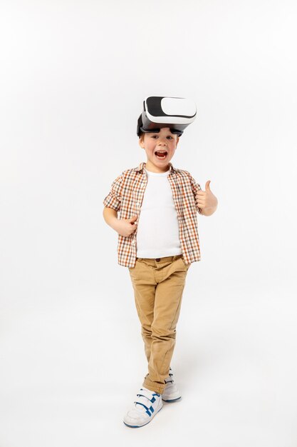Blij en benieuwd. Kleine jongen of kind in spijkerbroek en shirt met virtual reality headset bril geïsoleerd op witte studio achtergrond. Concept van geavanceerde technologie, videogames, innovatie.