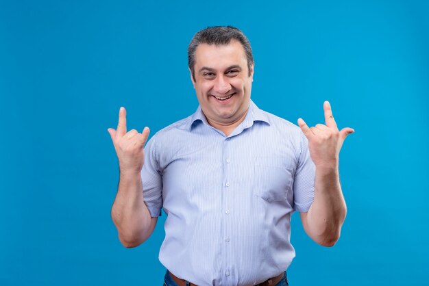 Blij en opgewonden man van middelbare leeftijd in blauw gestreept overhemd met rock gebaar met hand op een blauwe ruimte