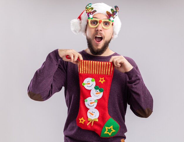 Blij en verrast jonge man in paarse trui en kerstmuts met grappige bril met een kerstsok kijken camera glimlachend staande op witte achtergrond
