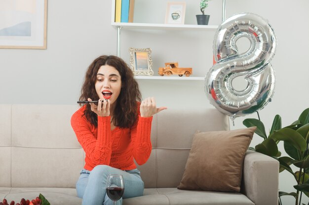 Blij en verrast jonge vrouw in casual kleding glimlachend vrolijk zittend op een bank met glas wijn praten op mobiele telefoon in lichte woonkamer vieren internationale vrouwendag 8 maart