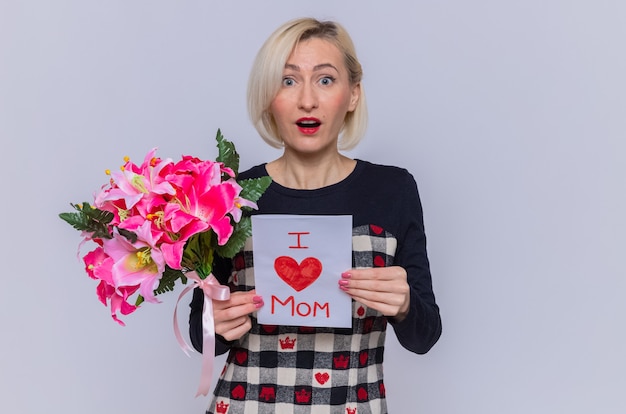 Blij en verrast jonge vrouw in een mooie jurk met wenskaart en boeket bloemen kijken naar voorzijde vieren moederdag staande over witte muur